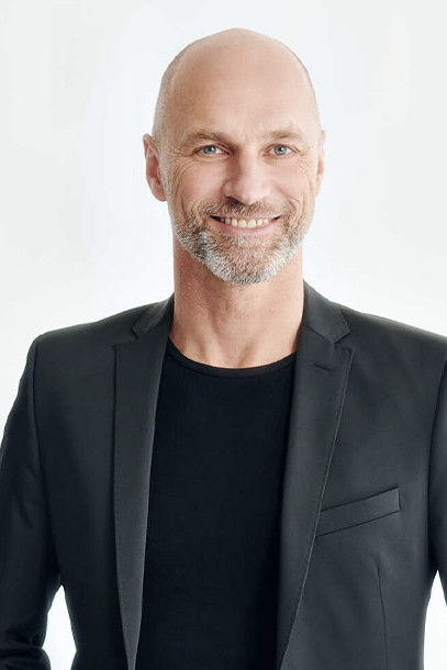 Lächelnder Mann mit kahlem Kopf und einem gräulichen Drei-Tage-Bart. Bekleidet mit einem schwarzen T-Shirt und einem grauen Jacket darüber- Jens Wohlfeil