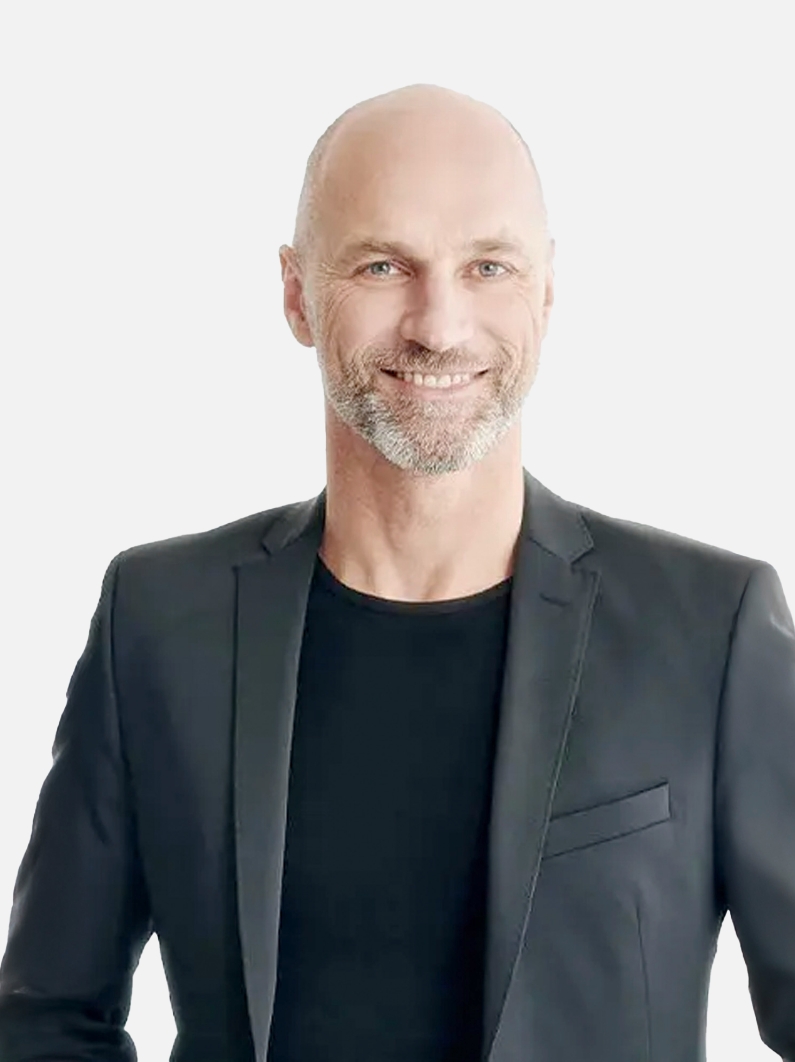 Lächelnder Mann mit kahlem Kopf und einem gräulichen Drei-Tage-Bart. Bekleidet mit einem schwarzen T-Shirt und einem grauen Jacket darüber- Jens Wohlfeil
